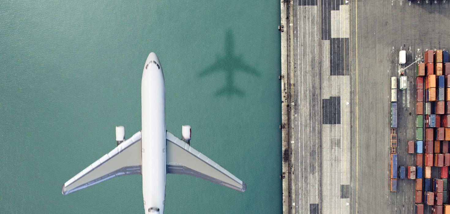 Bird's eye view of an airplane flying over water next to a shipping dock.