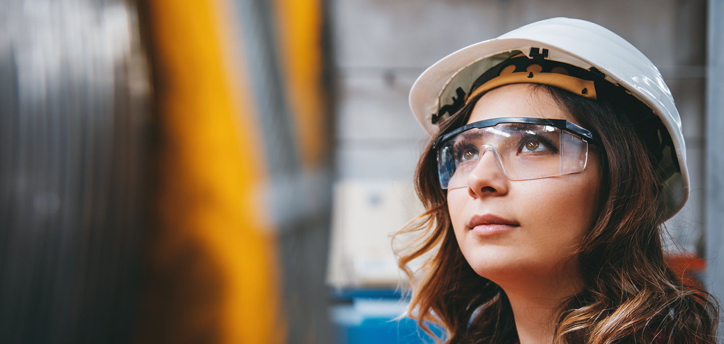 Woman in construction hat and safety glasses looking up inside a factory.
