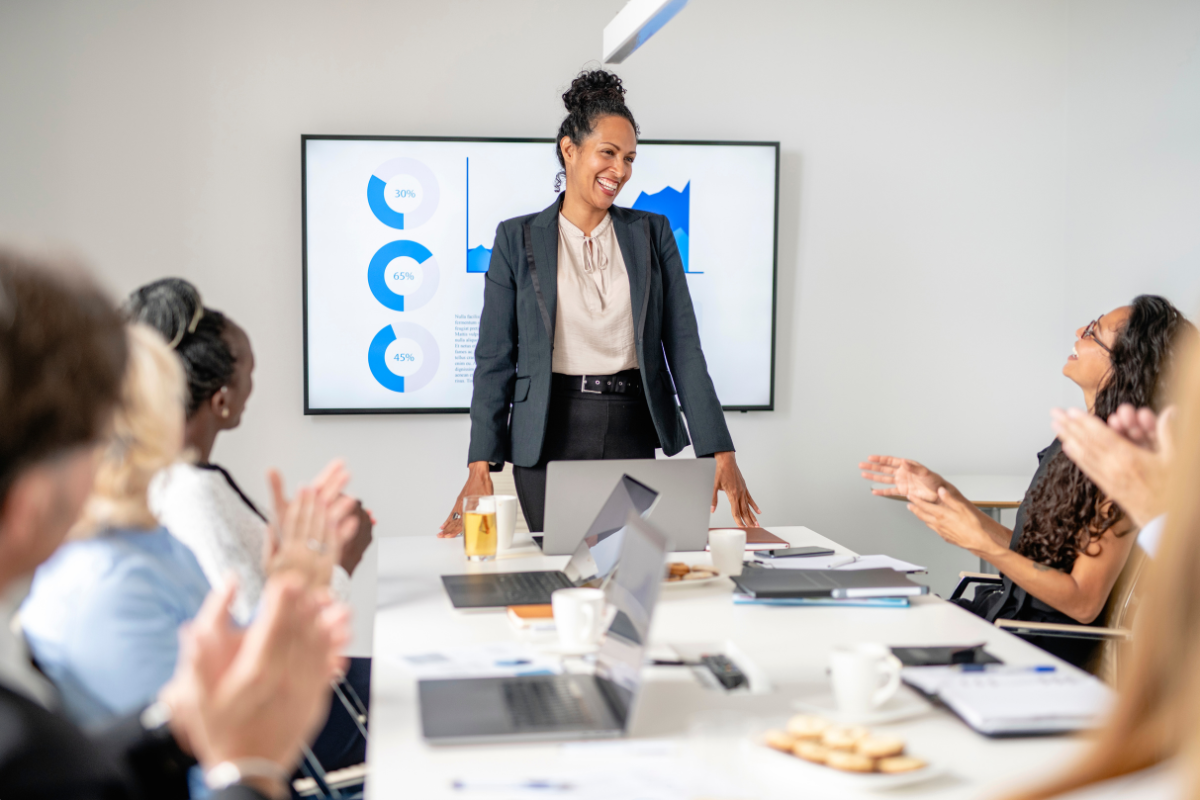A woman stands in front of a table filled with workers, she is leading a presentation.