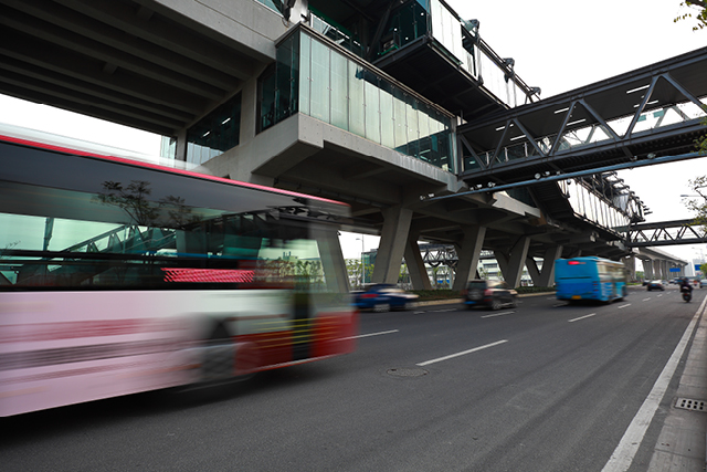 Blurred buses and vehicles move along a city roadway beneath a large elevated transit station and pedestrian walkway.